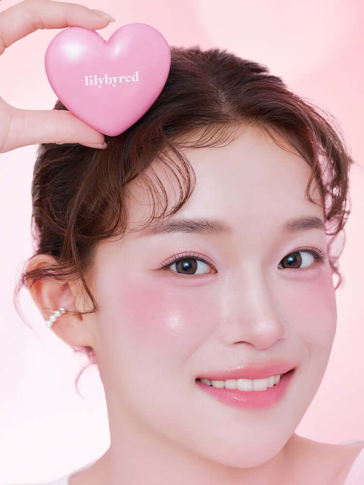 A smiling model holding a pink heart-shaped LILYBYRED Luv Beam Cheek Balm, showcasing a dewy glow on her cheeks.