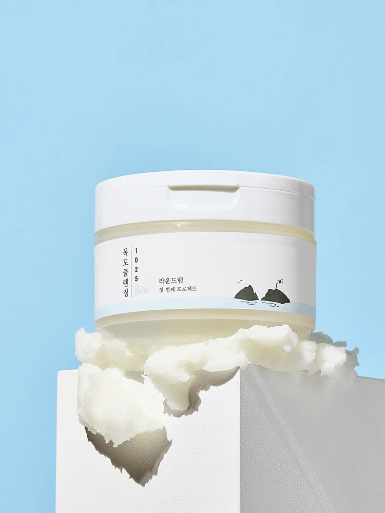 ROUND LAB 1025 Dokdo Cleansing Balm 100ml on a pedestal surrounded by a creamy texture against a blue background.